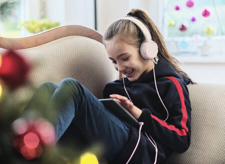 Fröhliches Mädchen, das im Urlaub auf dem Sofa im Wohnzimmer sitzt und Radio auf einem digitalen Tablet hört - DIKF00546