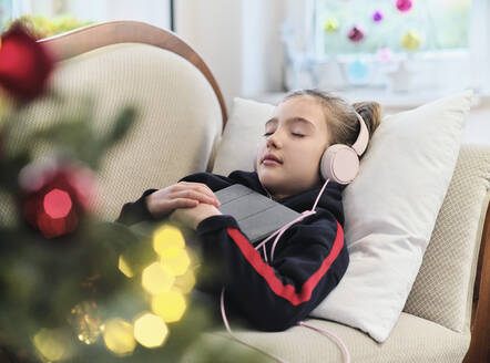 Entspanntes Mädchen, das auf dem Sofa im Wohnzimmer liegend Musik auf einem digitalen Tablet hört, während es Weihnachten feiert - DIKF00547