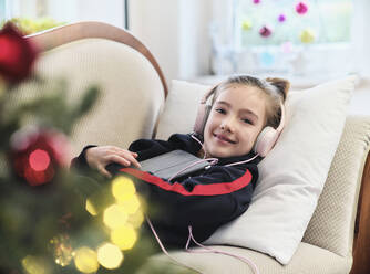 Lächelndes Mädchen mit digitalem Tablet, das über Kopfhörer Musik hört, während es auf dem Sofa zu Hause im Urlaub liegt - DIKF00548