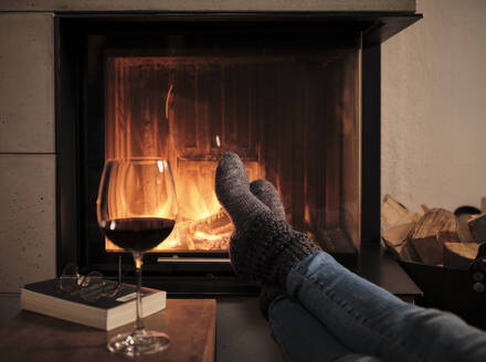 Woman wearing woolen socks resting feet on table by fireplace at home during winter - DIKF00549