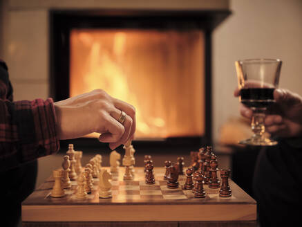 Reifer Mann mit Weinglas, während Frau am Kamin im Wohnzimmer Schach spielt - DIKF00552