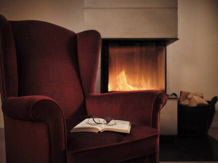 Eyeglasses with open book on maroon wingback chair by fireplace at home - DIKF00553