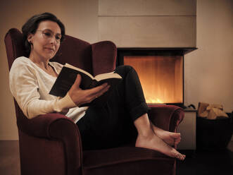 Mature woman reading book while sitting on armchair by fireplace at home - DIKF00555