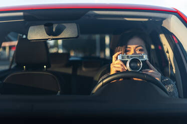 Junge Frau mit Vintage-Kamera im Auto durch die Windschutzscheibe an einem sonnigen Tag gesehen - JMPF00869