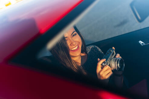 Lächelnde junge Frau mit Vintage-Kamera im Auto durch die Windschutzscheibe an einem sonnigen Tag gesehen - JMPF00871