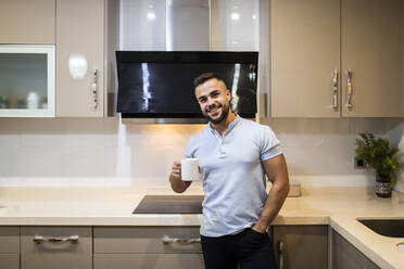 Hübscher junger Mann mit Händen in den Taschen, der eine Kaffeetasse hält, während er in der Küche steht - MIMFF00514