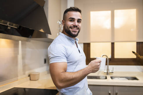 Muskulöser junger Mann lächelt und hält eine Kaffeetasse in der Küche - MIMFF00516