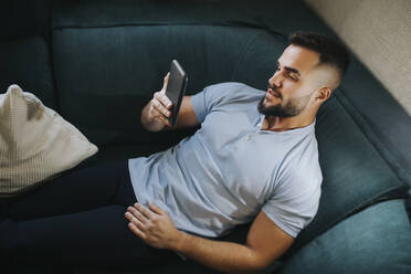 Handsome man using smart phone while lying on sofa at home - MIMFF00518