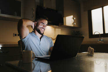 Lächelnder Geschäftsmann mit Händen hinter dem Kopf bei der Arbeit am Laptop am Esstisch zu Hause - MIMFF00520