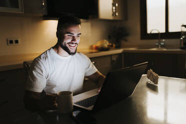 Bärtiger männlicher Unternehmer mit Kaffeetasse bei der Arbeit am Laptop am Esstisch in der Küche - MIMFF00521