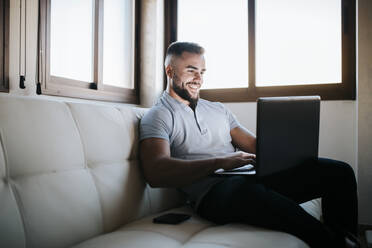 Lächelnder Geschäftsmann arbeitet am Laptop, während er zu Hause auf dem Sofa sitzt - MIMFF00524