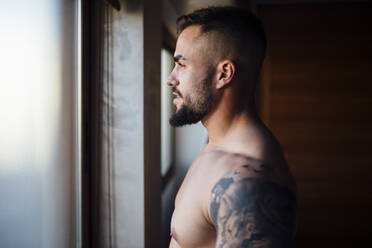Mann ohne Hemd mit Tätowierung schaut durch das Fenster zu Hause - MIMFF00527