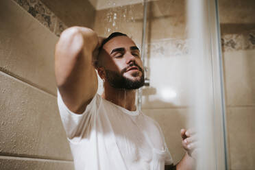 Young man in t-shirt with hand in hair taking shower in bathroom at home - MIMFF00531