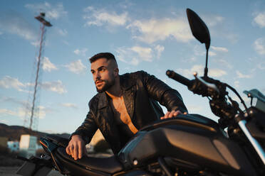 Gut aussehender Mann mit Lederjacke, der sich an ein Motorrad lehnt und an einem sonnigen Tag wegschaut - MIMFF00535