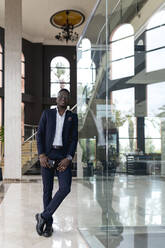 Young businessman leaning on glass wall in hotel lobby - EGAF01599