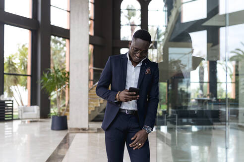 Smiling male entrepreneur using smart phone while leaning on glass wall in hotel lobby - EGAF01600