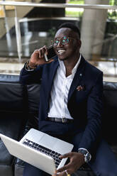 Lächelnder junger männlicher Unternehmer, der in der Hotellobby mit einem Laptop auf einem Smartphone spricht - EGAF01607