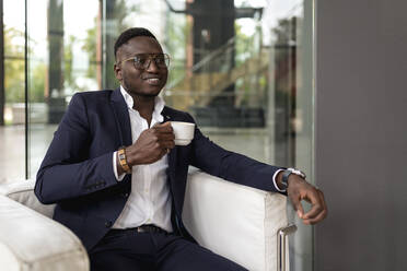 Young businessman holding coffee cup while day dreaming in hotel lobby - EGAF01613