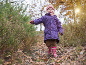 Smiling cute girl in purple jacket walking amidst plants - LAF02654