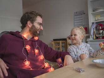 Lächelnder Vater mit Lichterkette um den Hals, der seine Tochter beim Essen in der Küche beobachtet - LAF02655