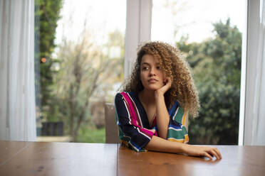 Nachdenkliche junge Frau sitzt am Tisch vor dem Fenster im Wohnzimmer - AXHF00120
