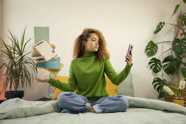 Young woman taking selfie through smart phone while tossing books in bedroom - AXHF00133