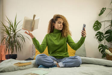 Young woman taking selfie on smart phone while books falling on hand in bedroom - AXHF00134