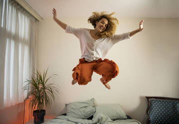 Cheerful young woman with arms raised jumping over bed in bedroom - AXHF00136