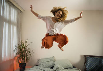 Fröhliche junge Frau mit erhobenen Armen, die über das Bett im Schlafzimmer springt - AXHF00136