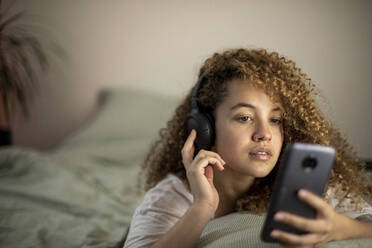 Young woman lying on bed while using smart phone and listening to music at home - AXHF00139