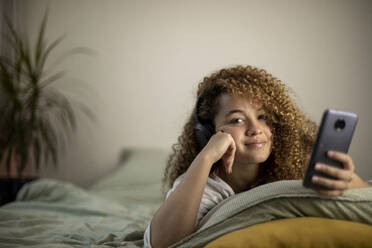 Smiling young woman with smart phone lying on bed in bedroom - AXHF00140