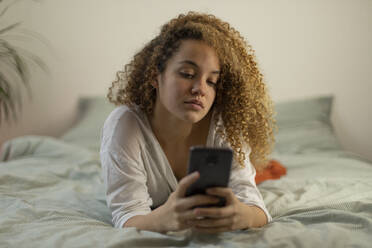 Young woman text messaging through mobile phone while lying on bed in bedroom - AXHF00142