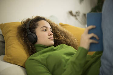 Junge Frau mit Buch auf dem Bett liegend, während sie zu Hause über Kopfhörer Musik hört - AXHF00143