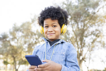 Lächelnder Junge mit Kopfhörern, der ein Mobiltelefon benutzt, während er im Park steht - JCCMF00958