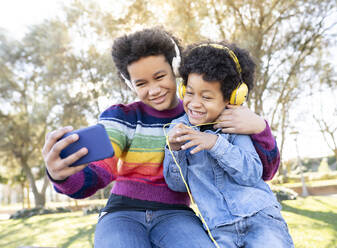 Sibling wearing headphones smiling while taking selfie through mobile phone while sitting at park - JCCMF00960