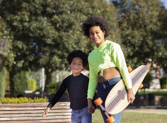 Mädchen hält Skateboard und die Hand ihres Bruders beim Spaziergang im Park - JCCMF00992