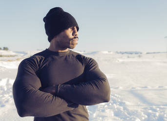 Sportsman with arms crossed looking away while standing in snow during sunny day - JCCMF01005