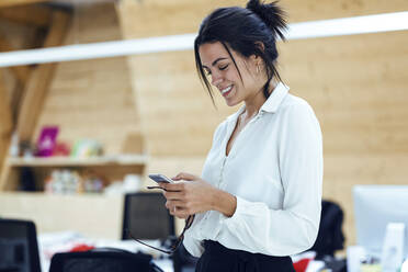 Smiling female entrepreneur texting through mobile phone at office - JSRF01307