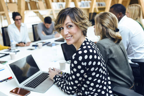 Lächelnde Geschäftsfrau sitzt mit Kollegen am Konferenztisch im Sitzungssaal während einer Sitzung - JSRF01319