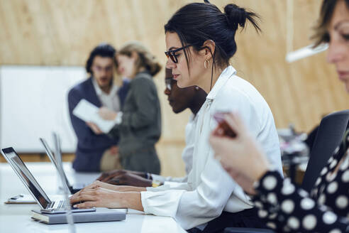 Geschäftsfrau, die einen Laptop benutzt, während sie inmitten von Kollegen in einem Coworking-Büro sitzt - JSRF01327
