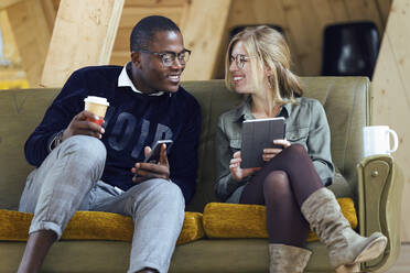 Smiling male and female professionals discussing over digital tablet while sitting on sofa at coworking office - JSRF01329