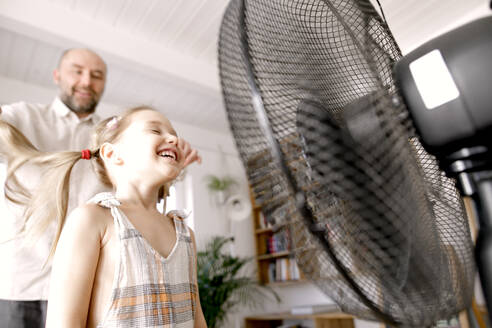 Girl in front of electric fan while father standing at home - KMKF01480