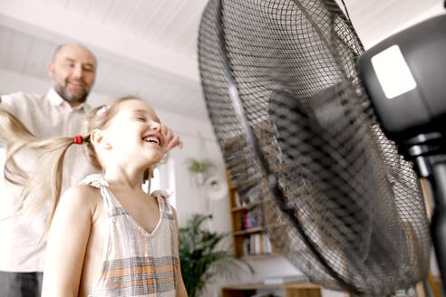 Mädchen vor einem elektrischen Ventilator, während der Vater zu Hause steht - KMKF01480