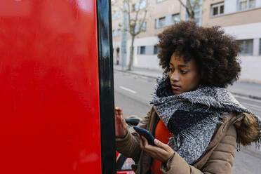 Afro woman with smart phone buying ticket from machine during winter - XLGF01047