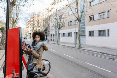 Young woman looking away while standing by ticket machine at bicycle parking station in city - XLGF01048