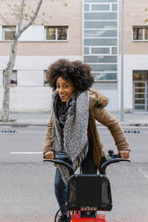 Cheerful Afro woman sitting on bicycle against building on street - XLGF01049