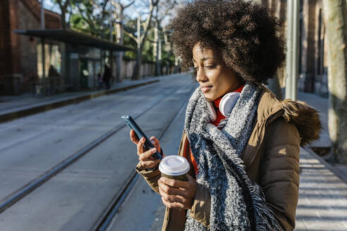 Junge Frau mit Kaffeetasse und Mobiltelefon auf der Straße an einem sonnigen Tag - XLGF01057