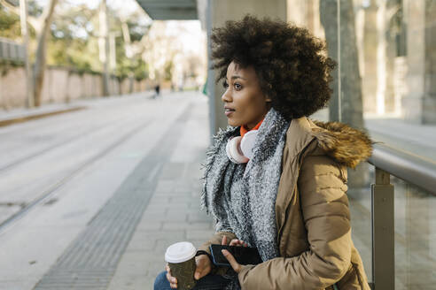 Afro-Frau mit Smartphone und Einweg-Kaffeebecher nachdenklich am Busbahnhof im Winter - XLGF01058