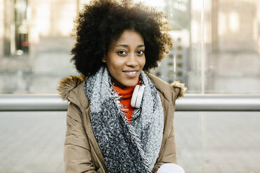 Smiling woman in warm clothing with headphones against glass at bus stop - XLGF01059