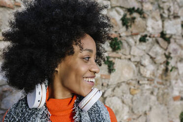 Happy Afro woman with headphones looking away against stone wall - XLGF01064